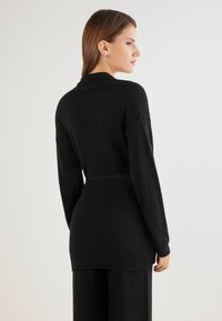 Cardigan negro de punto con mangas largas y un cinturón. Presenta un cuello chal, una tela texturizada y un ajuste relajado que cae por debajo de las caderas.