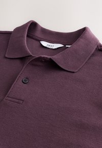 Polo bordeaux en tissu texturé avec col plat, un seul bouton noir et une petite poche poitrine, étiqueté "NEXT REGULAR FIT."