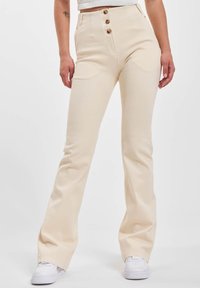 Kvinne i høy midje, kremfargede, vide bukser med tre knapper og hvite sneakers, som står mot en ensfarget bakgrunn.