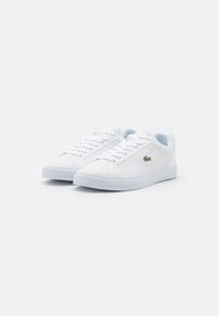 White sneakers made of synthetic material, featuring a smooth texture, round toe, flat sole, and a small green logo accent.