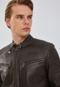 Giacca in pelle marrone con vestibilità slim, dotata di alto colletto, cuciture diagonali sulle spalle e due tasche frontali con zip.