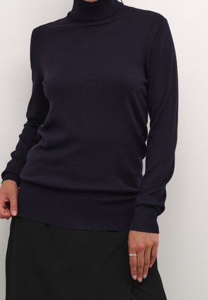 Person trägt einen dunkelblauen Rollkragenpullover mit langen Ärmeln und einen schwarzen Rock und steht vor einer einfarbigen hellen Wand.