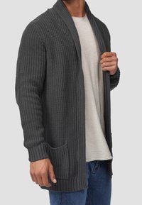 Hombre vistiendo un cárdigan de punto texturizado gris oscuro sobre una camisa blanca y jeans azules, sosteniendo el cárdigan abierto con una mano.