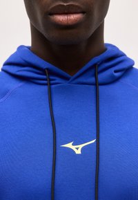 Κοντινό πλάνο ενός ατόμου που φοράει μπλε φούτερ με κουκούλα, μαύρα κορδόνια και λευκό λογότυπο Mizuno στο στήθος.