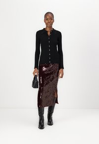 Schwarze gerippte Strickjacke mit goldenen Knöpfen, kombiniert mit einem bordeauxfarbenen Paillettenrock mit einem Seitenschlitz und schwarzen kniehohen Stiefeln.