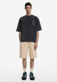 Camiseta negra desgastada y oversize con logo blanco, pantalones cortos beige y zapatillas oscuras con acentos en verde azulado. Modelo de pie contra un fondo blanco.
