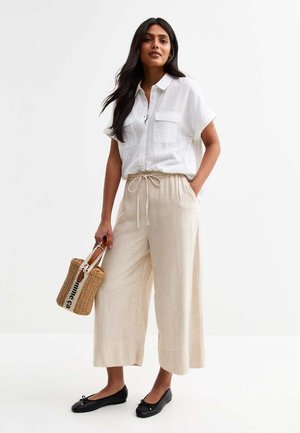 Chemise blanche légère à manches courtes avec boutons, pantalon large beige avec cordon de serrage, ballerines noires et un petit sac en osier.