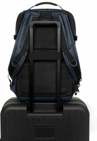 Mochila azul marino con respaldo de malla, correas ajustables y funda trasera para trolley. Colocada sobre una maleta rígida negra con estrías verticales.