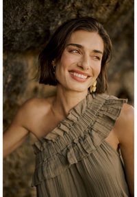 Ühe õlavarrega kleit tan värvi kangast, millel on vertikaalsed plisseeritud detailid ja ripskoes serv. Taustal on tekstureeritud kivi, mis tõhustab looduslikku keskkonda.