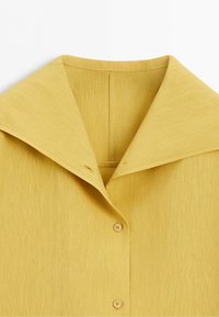 Yellow shirt with a wide pointed collar and two visible buttons on a smooth fabric background.