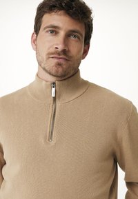 Beige svetr s vysokým límcem a čtvrtinovým zipem. Rybářský vzor a slim fit. Zdobí ho malé zlaté logo na hrudi.