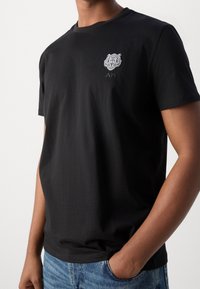 Black cotton t-shirt with short sleeves, featuring a white embroidered tiger design on the chest and a subtle brand logo below.