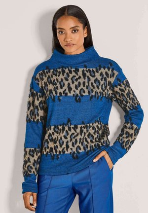 Frau in einem blauen Pullover mit beigen und schwarzen Leopardenmusterstreifen, kombiniert mit blauen Hosen, steht mit einer Hand in der Tasche.