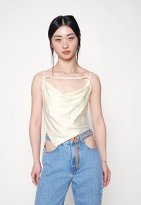 Top de cetim com decote cowl em amarelo pálido, apresenta alças finas e uma bainha assimétrica. Combinado com jeans de denim azul de cintura alta.