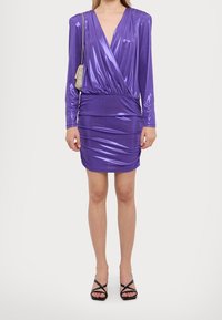 Fialové šaty s dlouhými rukávy a překříženým designem, vyrobené z elastického lesklého materiálu, s přiléhavým siluetou a řasením v pase. Černé sandály na podpatku.