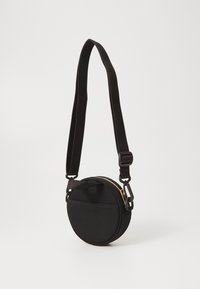 Black circular bag with a textured fabric, gold zipper, adjustable strap, and a small front pocket. Simple, minimalist design.