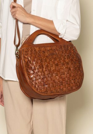 Frau, die eine braune geflochtene Ledertasche in runder Form und ein beiges Outfit vor einem neutralen Hintergrund hält.