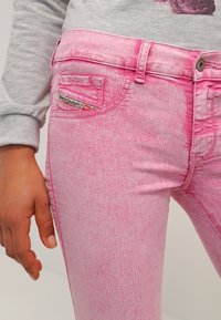 Lähikuva henkilöstä, jolla on päällä vaaleanpunaiset happovärjätyt farkut ja harmaa pitkähihainen paita, jossa on kukkakuosi, käsi rentona sivulla.