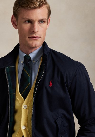 Bărbat purtând o jachetă bleumarin cu logo roșu peste un cardigan galben, cămașă albastră cu nasturi și cravată verde cu dungi, uitându-se în lateral pe un fundal simplu.