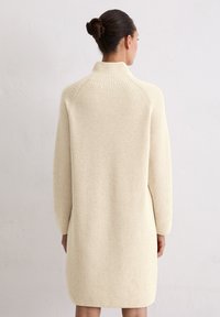 Kremowa dzianinowa sukienka z wysokim kołnierzem, długimi rękawami i fakturowanym wzorem, sięgająca tuż nad kolana.