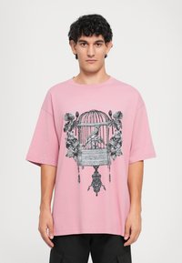 Ροζ βαμβακερό T-shirt με κοντά μανίκια, που διαθέτει μαύρη εκτύπωση ενός κλουβιού που είναι περιτριγυρισμένο από λουλούδια και διακοσμητικές λεπτομέρειες.