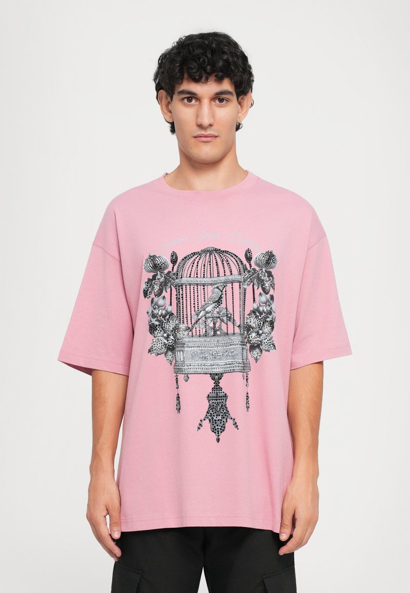 Ροζ βαμβακερό T-shirt με κοντά μανίκια, που διαθέτει μαύρη εκτύπωση ενός κλουβιού που είναι περιτριγυρισμένο από λουλούδια και διακοσμητικές λεπτομέρειες.