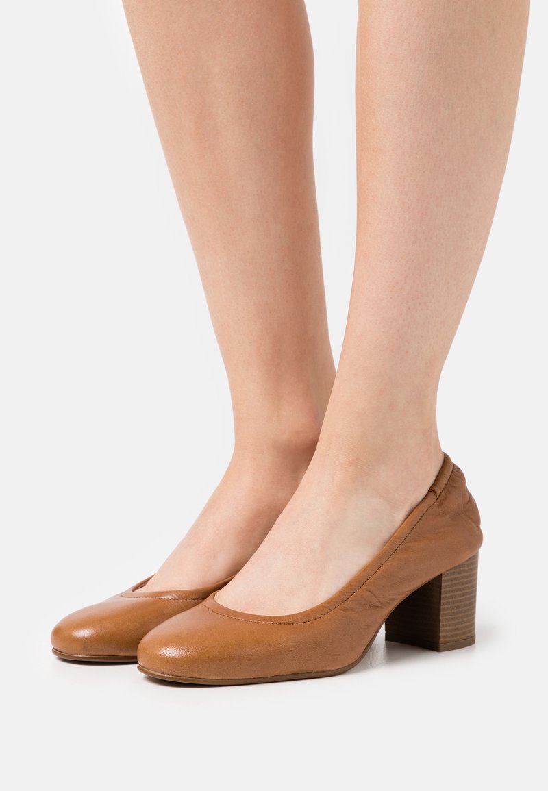 Jambes portant des chaussures en cuir marron clair à talons blocs bas avec bouts arrondis, sur un fond clair uni.