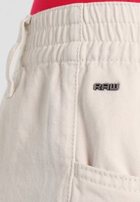 Beiga byxor med elastisk midja, snörningsdetalj och en liten "RAW"-logotyp på tyget. Strukturerad bomullsmaterial och sidoficka.