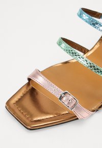 Sandale à plusieurs brides avec un design à bout carré. Présente des brides métalliques rose, verte et bleue, une base en bronze et un détail de boucle en argent.