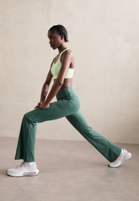 Pantaloni da yoga verdi e un reggiseno sportivo verde chiaro sono indossati con sneaker bianche. I pantaloni hanno un design svasato e una texture liscia.
