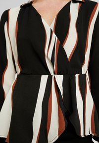 Blusa a righe nera, bianca e ruggine con design avvolgente, maniche lunghe, colletto e vita a peplo. Tessuto testurizzato con un lieve lucido.