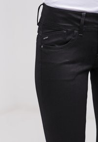 Jeans de denim negro con corte ajustado, con bolsillos frontales, costuras sutiles y un acento de logotipo en la cinturilla. Textura suave, diseño minimalista.