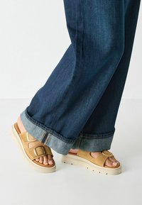 Beige suede sandals with adjustable buckles and a thick, cream-colored sole. Worn with dark denim jeans, featuring a cuffed hem.