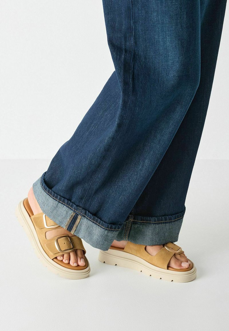 Beige suede sandals with adjustable buckles and a thick, cream-colored sole. Worn with dark denim jeans, featuring a cuffed hem.