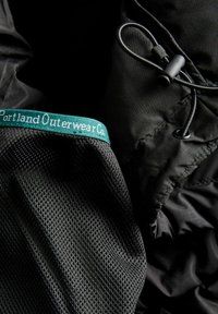 Černé svrchní oblečení s texturovanou mřížkovou podšívkou, s zelenou nálepkou s nápisem "Portland Outerwear Co." a černým nastavitelným šňůrkovým uzávěrem.