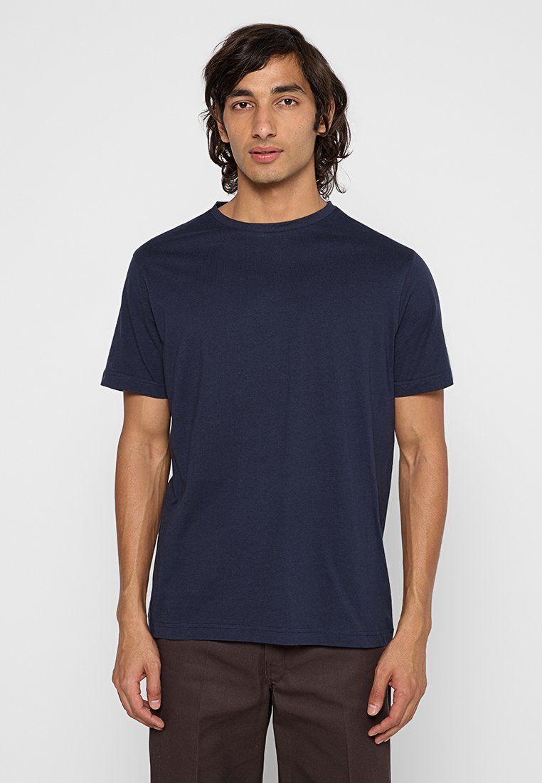 Mann in einem schlichten, navyblauen T-Shirt mit kurzen Ärmeln und dunkelbraunen Hosen, der vor einem schlichten, hellgrauen Hintergrund steht.