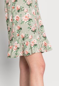 Vestido floral em tecido verde claro, com rosas cor-de-rosa e brancas, bainha com volantes e corte a meio da altura. Textura suave, design ajustado.