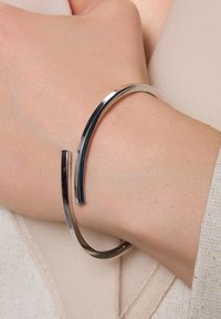 Srebrna metalowa bransoletka na nadgarstek o zakrzywionym kształcie i przerwie, prezentująca polerowane wykończenie oraz elegancki, minimalistyczny styl.