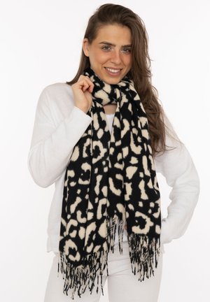 Lächelnde Frau mit langen braunen Haaren, die ein weißes Oberteil und eine weiße Hose trägt und einen schwarz-weiß gemusterten Schal mit Fransenenden hält.