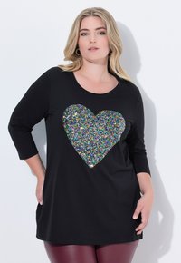 Tunique noire à manches longues avec un grand cœur en sequins multicolores au centre, fabriquée en tissu doux offrant un ajustement confortable.