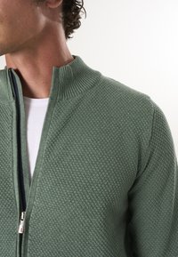 Suéter verde con textura, con cierre frontal y cuello acanalado. El material presenta un patrón elevado, visible parcialmente en el cuello y el hombro del modelo.