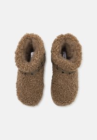 Bruna fluffiga tofflor med ett texturerat yttermaterial, rund tå och en mjuk fleeceinredning. Har ett brunt band med svarta cirkulära detaljer.