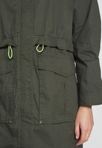 Chaqueta ligera de color verde oliva con dos bolsillos delanteros, cordón ajustable en la cintura y detalles en verde neón en el cordón y las costuras.