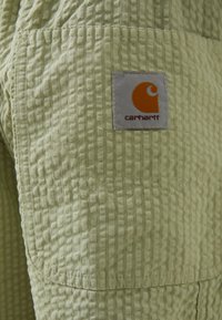 Pantalones de tela texturizada verde claro con un patrón de cuadrícula, que presentan un destacado parche de logo naranja cosido en el bolsillo lateral.