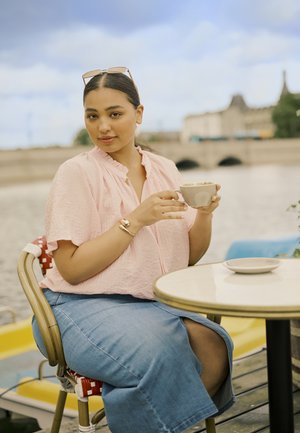 Blusa rosa chiaro, texutata, con scollatura arricciata; gonna di jeans. Tiene in mano una tazza chiara, seduta a un tavolo rotondo di un caffè vicino all'acqua.