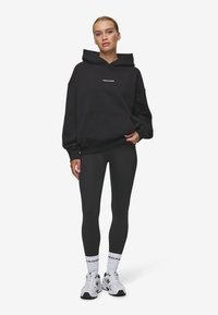 Zwarte oversized hoodie met een voorzak en logo, gecombineerd met zwarte leggings en witte sneakers met zwarte accenten en branding.
