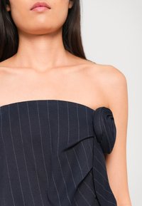 Femme portant un top bustier bleu marine foncé à fines rayures avec un grand détail de rose en tissu sur un côté, sur un fond clair uni.