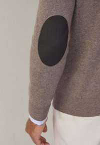 Ruskea neulepaita, jossa on musta kyynärpääpaikka. Ribbikujat, joissa on vaaleansininen ja valkoinen raidoitus alla olevassa paidassa.