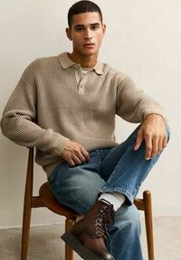 Młody mężczyzna w beżowym prążkowanym swetrze, niebieskich dżinsach i brązowych sznurowanych butach, siedzący ze skrzyżowanymi nogami na drewnianym krześle na jednolitym tle.