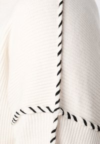 Wit gebreid stof met zwart gedraaid koord langs de randen en naden, met een gedetailleerde textuur en patroon van het materiaal.